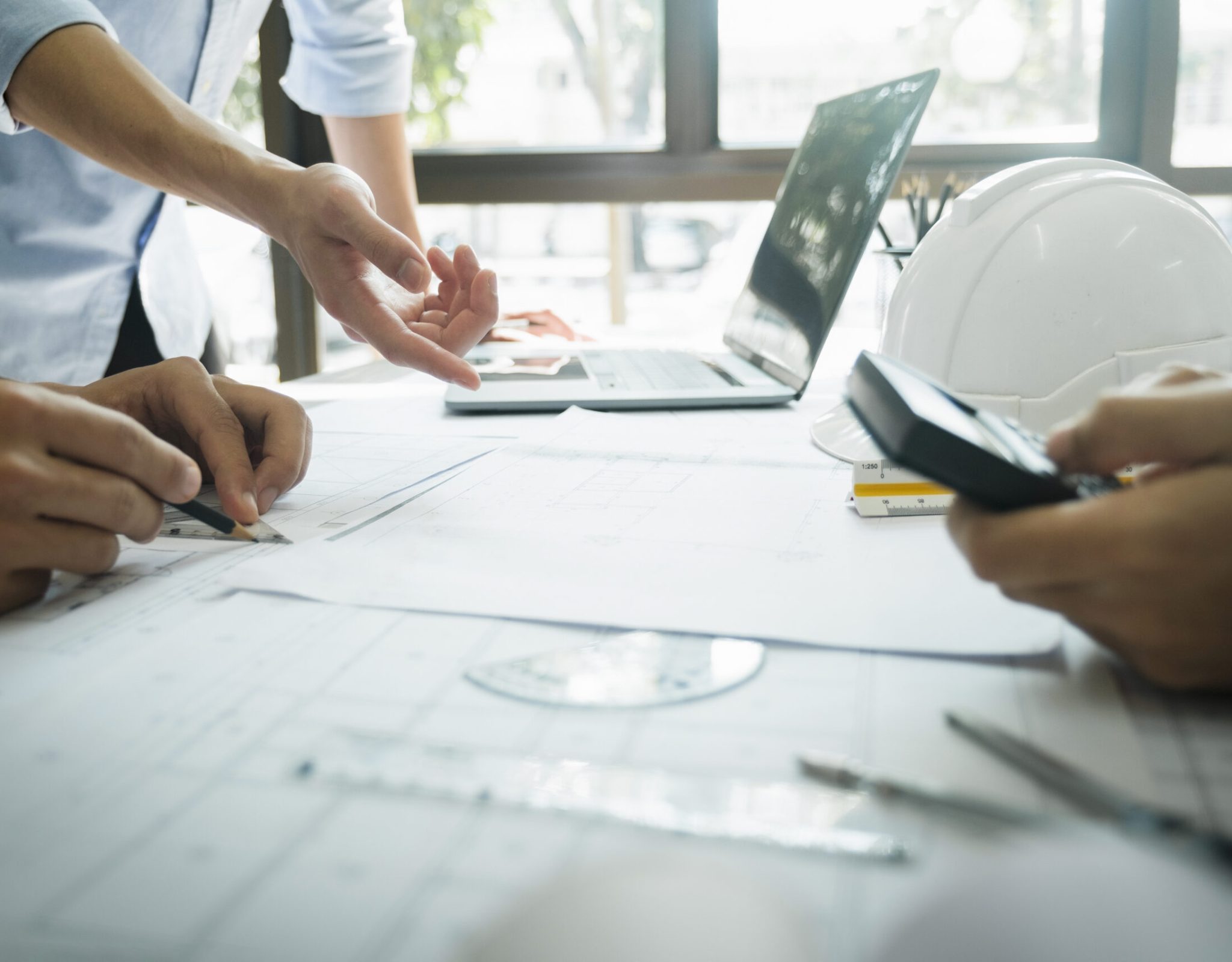 Close-up hands shot of three engineer, developers, or architects working together, drawing graphic design of construction project, and discussing for improvement  at desk with papers, compass, helmet, calculator, rulers, and laptop. Engineering and Teamwork concept.
